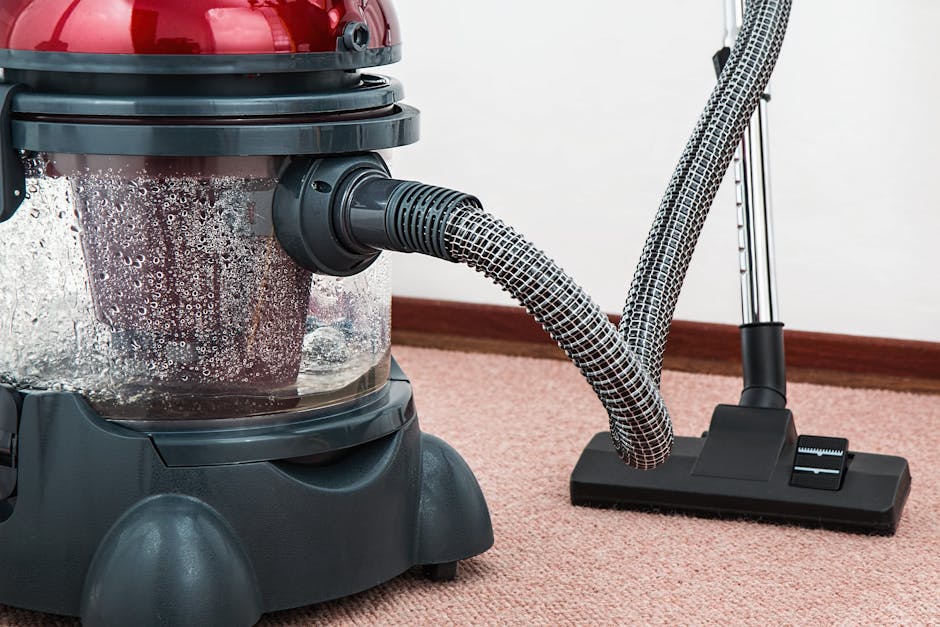 A professional deep cleaning process underway inside a residential living room, where a cleaner dressed in a white protective suit, face mask, gloves, and shoe covers is using a vacuum or extraction t