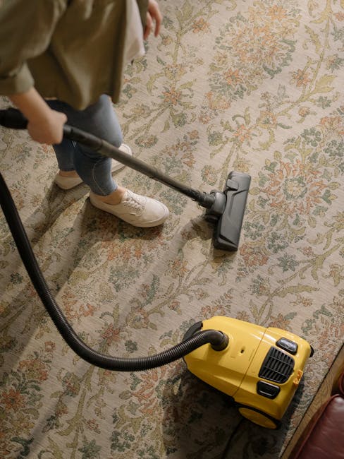 A person, wearing white sneakers and beige pants, is using a vacuum cleaner on a patterned area rug with floral motifs in shades of beige, green, and rust. The vacuum cleaner is a yellow canister mode