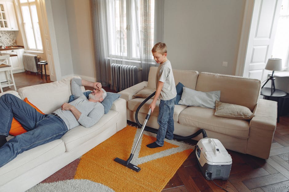 A person, wearing white sneakers and beige pants, is using a vacuum cleaner on a patterned area rug with floral motifs in shades of beige, green, and rust. The vacuum cleaner is a yellow canister mode